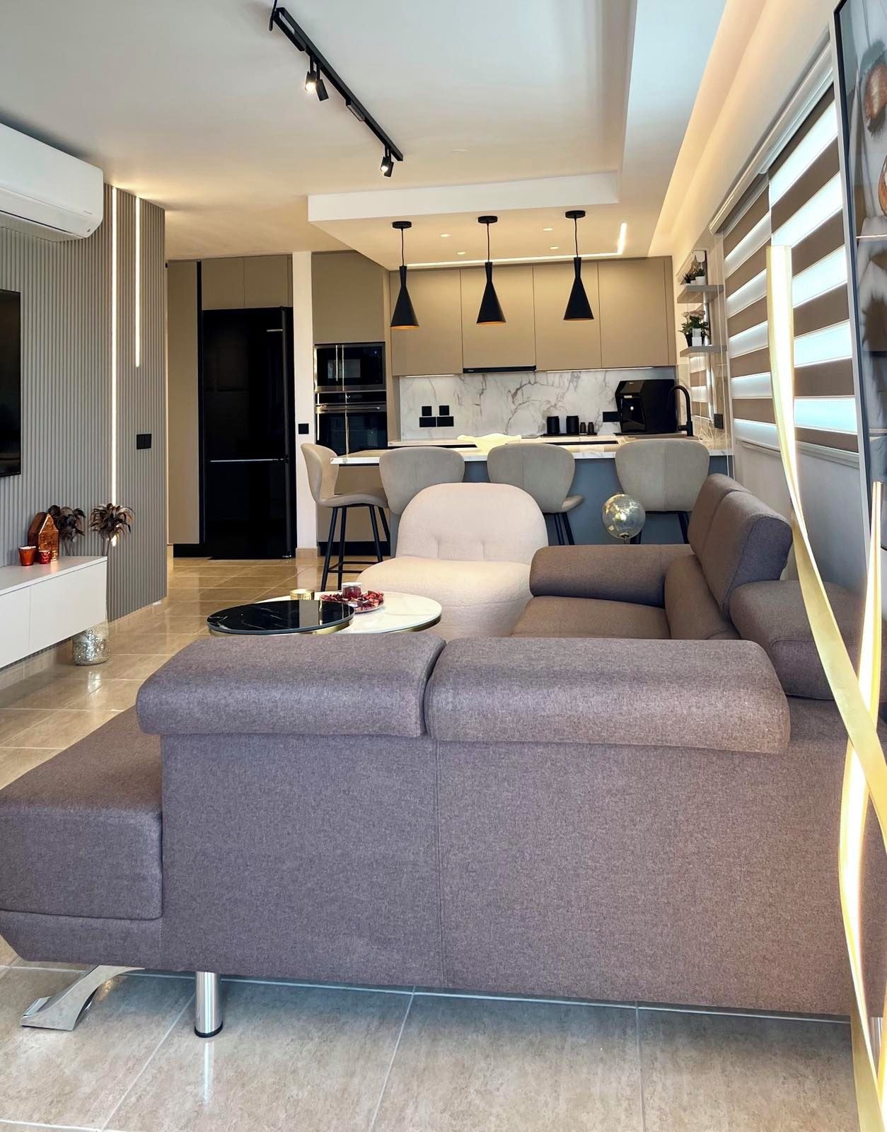 Una sala moderna con un sofá seccional, un comedor con taburetes de bar y una cocina con iluminación colgante.