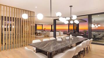 Un espace repas moderne avec une grande table en marbre, des chaises blanches et une vue sur le coucher de soleil à travers de grandes fenêtres.