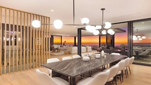 Een moderne eetruimte met een grote marmeren tafel, witte stoelen en uitzicht op de ondergaande zon door grote ramen.
