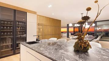 Une cuisine moderne dotée d’un plan de travail en marbre, d’un meuble à vin intégré et offrant une vue sur le coucher de soleil grâce à de grandes fenêtres.