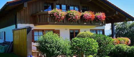 Una casa de madera de dos pisos con un balcón decorado con flores de colores, un césped cuidado y un pequeño cobertizo.