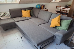 A grey sectional sofa with yellow and green cushions, a small table with a globe and books, and a dining area with a table and chairs. - Holiday apartment Mosselfischer (Mehring)
