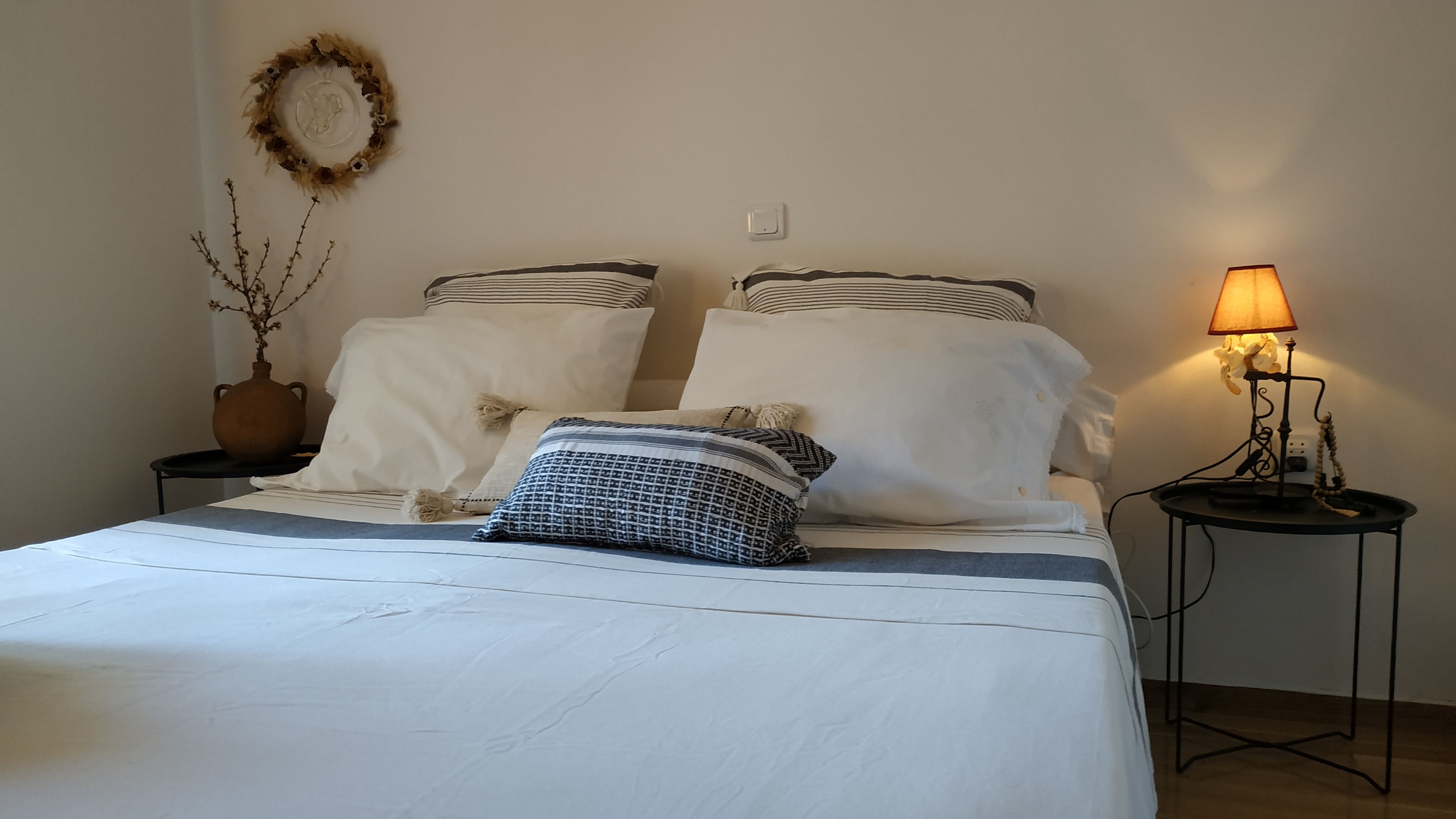 A neatly made bed with white bedding and striped pillows, a vase with branches on a side table, and a lamp on another table.