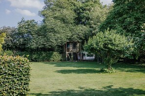 Un jardín con una estructura de madera, posiblemente un pabellón, rodeado de abundante vegetación y un césped cuidado.