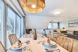 A dining area with a table set for four, surrounded by wicker chairs, a bottle of Manz wine, and a view of the living area. - Holiday apartment in Sylt with 2 bedrooms (Westerland)