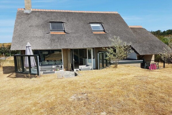 Une maison avec un toit en pente, deux lucarnes, une entrée avec une porte vitrée et un petit espace extérieur aménagé pour s’asseoir.