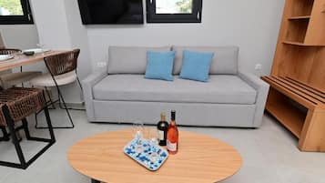A modern living room with a grey sofa, two blue cushions, a wooden coffee table with wine glasses and a bottle, and a TV mounted above a window.