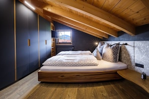 Ein Schlafzimmer mit schrägem Dach, einem Bett mit gemusterten Betttextilien und Blick ins Freie durch ein Fenster.