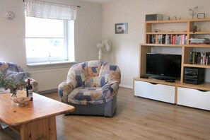 A living room with a sofa, a television on a stand, a bookshelf, and a window with curtains. - Holiday apartment with 1 bedroom (Garding Kspl.)