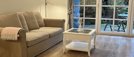 A beige sofa with a throw blanket, a white side table with a candle, and a large window with a view of greenery.