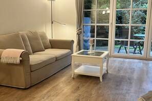 A beige sofa with a throw blanket, a white side table with a candle, and a large window with a view of greenery. - Holiday apartment with 1 bedroom (Sankt Peter-Ording)
