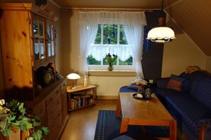A cozy living room with a wooden cabinet, a table with a lamp, a sofa, and a window with a view of greenery. - in Gretchen's house 2 (Südbrookmerland Ortsteil Moorhusen)