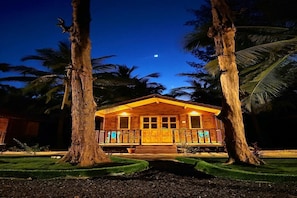 Una cabaña de madera con un porche, rodeada de palmeras y un cielo nocturno despejado.