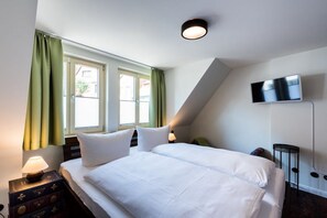 A hotel room with a bed, a television mounted on the wall, a nightstand with a lamp, and a window with curtains. - To the cheerful day laborer (Quedlinburg)