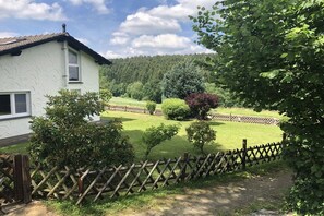 Ein Haus mit weißer Fassade, einem eingezäunten Garten und einem bewaldeten Hügel im Hintergrund.