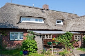 A traditional thatched-roof house with a brick exterior, a small window, and a garden area with a wheelbarrow and potted plants. - Thatched roof house - House 2 (Wittdün auf Amrum)