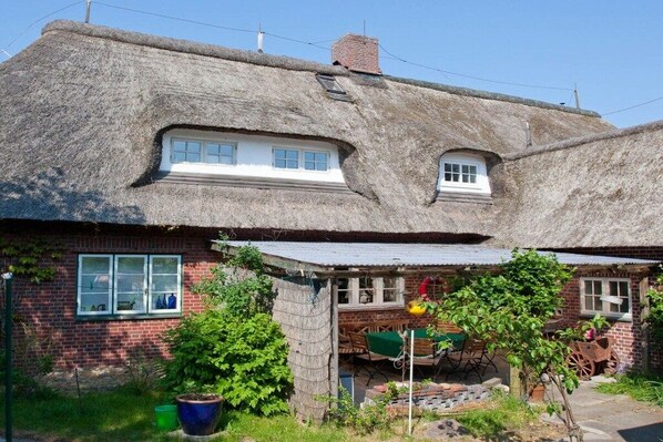 Ein traditionelles Haus mit Strohdach und Backsteinfassade, einem kleinen Fenster und einem Gartenbereich mit Schubkarre und Kübelpflanzen.