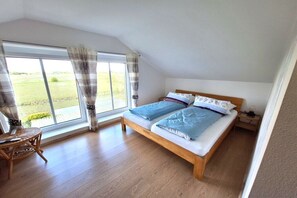 A bedroom with a wooden bed, a small bedside table, a chair, and a large window with curtains. - Holiday apartment with 2 bedrooms (Pellworm)