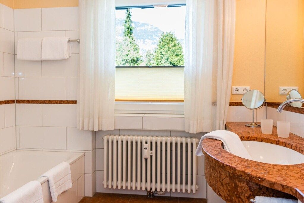 A bathroom with a bathtub, a sink with a mirror, and a window with a view of greenery.