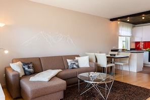 A modern living room with a brown sofa, a glass coffee table, and a dining area with white chairs. - Holiday apartment with 1 bedroom (Garmisch-Partenkirchen)