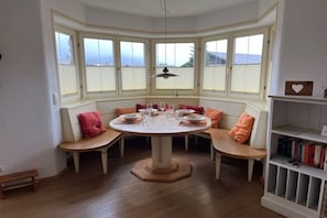 A round dining table with six wooden benches, a white pendant light, and a bookshelf with books. - Holiday apartment with 2 bedrooms (Garmisch-Partenkirchen)