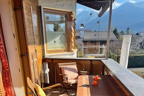 A wooden deck with a table and chairs, a window with blinds, and a view of a mountain landscape. - Holiday apartment with 2 bedrooms (Garmisch-Partenkirchen)