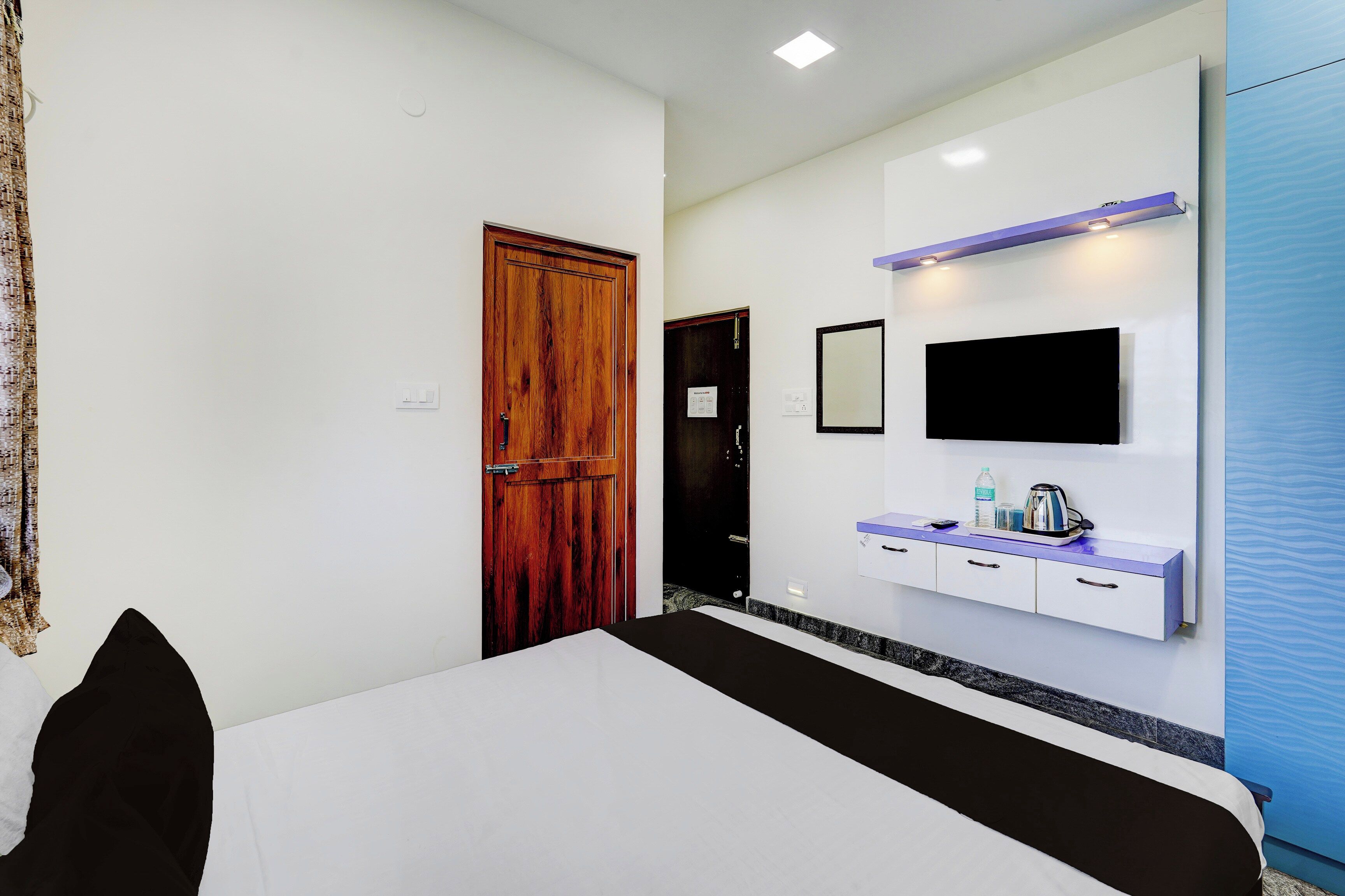 A hotel room with a bed, a wooden door, a flat-screen TV mounted on the wall, a small table with a kettle and glasses, and a bathroom door.