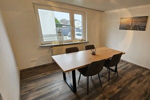 A dining area with a wooden table and chairs, a radiator below a window, and a cityscape painting on the wall. - Holiday apartment on the edge of the forest (Wassenberg)