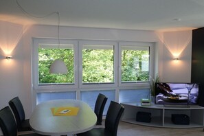 Ein modernes Wohnzimmer mit Esstisch, Stühlen, einem Flachbildfernseher und einem großen Fenster mit Blick auf Grünflächen.