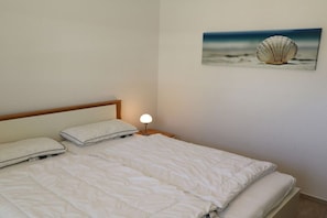Een netjes opgemaakt bed met wit beddengoed en kussens, een bedlampje en een lijst met een foto van een schelp aan de muur.