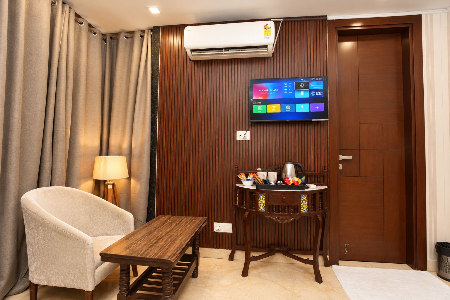 A hotel room with a wooden table, a cushioned chair, a television mounted on the wall, and a wooden cabinet with kitchen items.