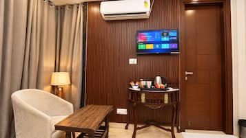 A hotel room with a wooden table, a cushioned chair, a television mounted on the wall, and a wooden cabinet with kitchen items.