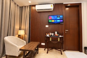 Habitación de hotel con una mesa de madera, una silla acolchada, un televisor en la pared y un mueble de madera con utensilios de cocina.