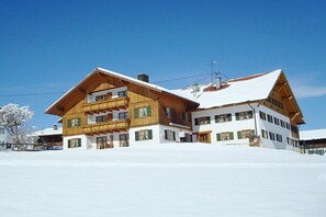 A large wooden chalet with multiple balconies and a snow-covered roof, situated in a snowy landscape. - 3 - at the Füssen equestrian center (Weißensee-Füssen)