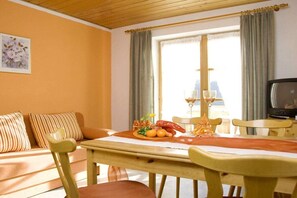 A room with a wooden ceiling, a sofa with striped cushions, a dining table with a fruit plate, a television, and a window with curtains. - 3 - at the Füssen equestrian center (Weißensee-Füssen)