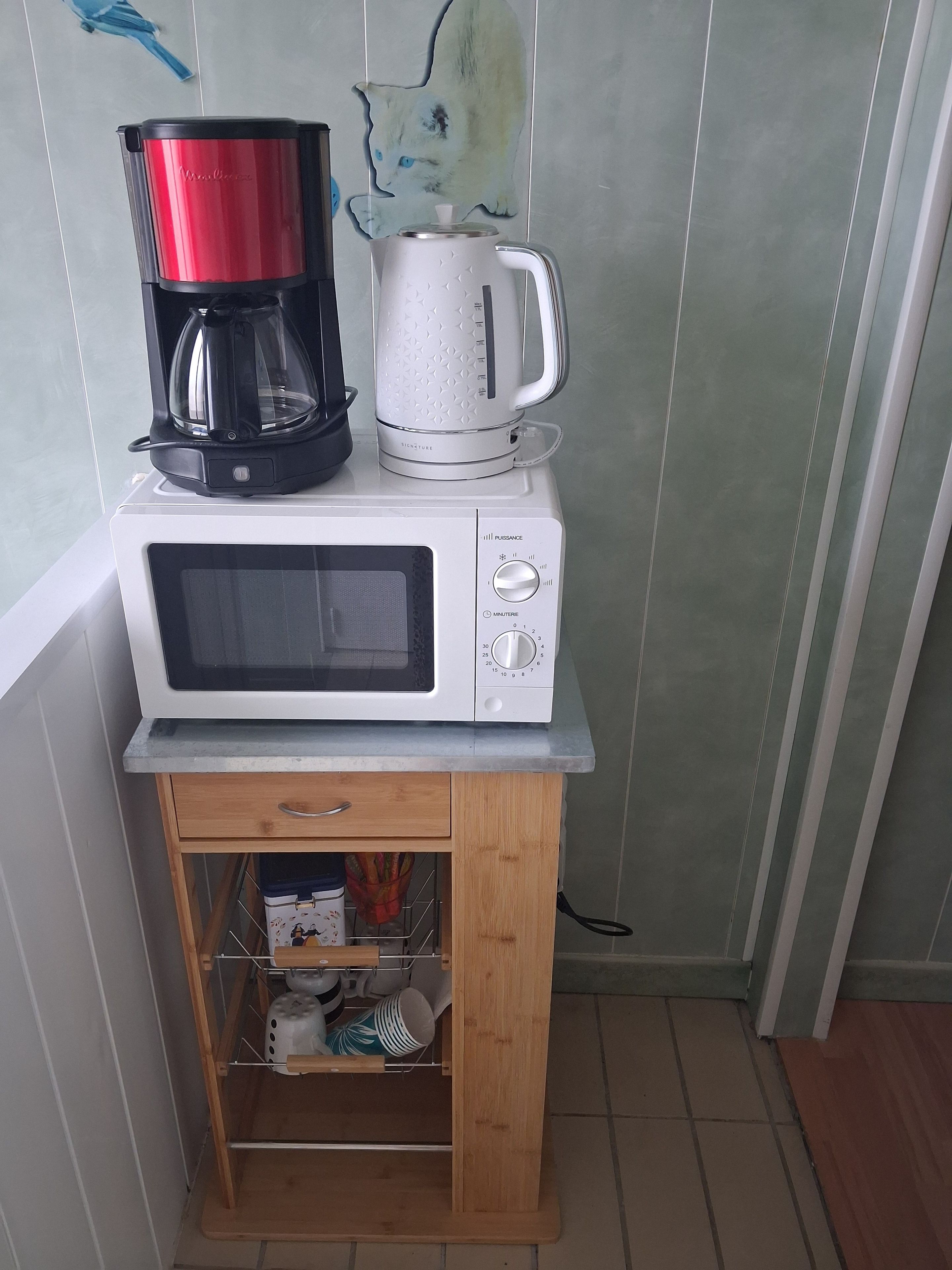 Cozinha compacta com micro-ondas, cafeteira e chaleira sobre um pequeno armário de madeira.