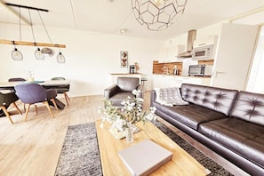 A modern living room with a leather sofa, a dining table with chairs, a coffee table with a vase of flowers, and a kitchen area with cabinets and appliances. - Holiday apartment with 1 bedroom (Buren\/Ameland)