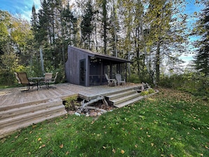 Una terraza de madera con mobiliario exterior, una pequeña construcción tipo cabaña y un fondo de bosque.