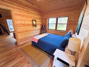 Un dormitorio de cabaña de madera con cama, mesita de noche, lámpara y ventana.