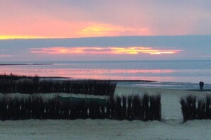 A beach scene with a sunset, a person standing on the sand, and vegetation in the foreground. - 2 in the periwinkle house (Cuxhaven-Duhnen)