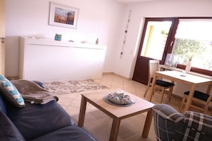 A living room with a sofa, a coffee table, a dining table with chairs, a radiator, and a window with a view. - 2 in the periwinkle house (Cuxhaven-Duhnen)