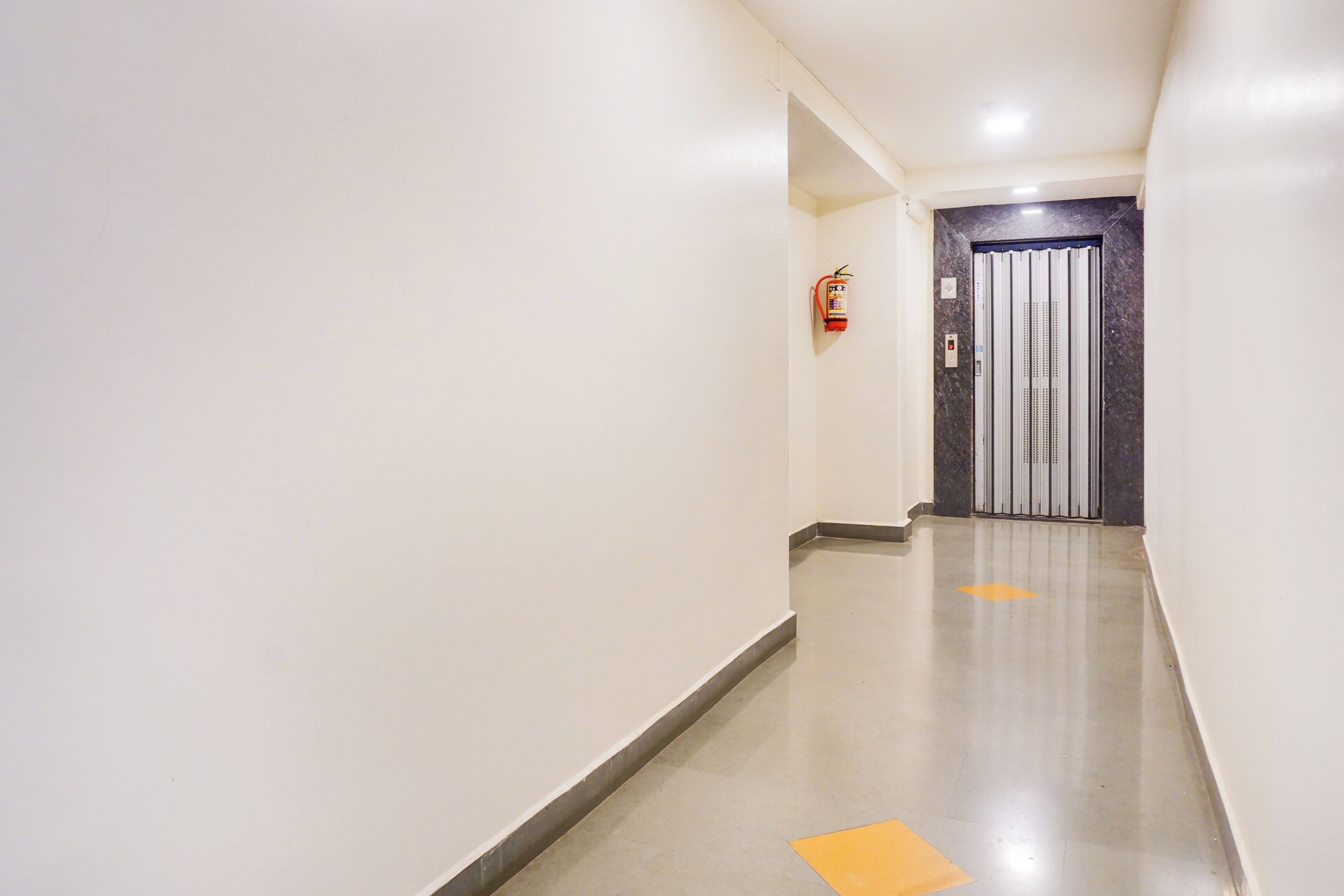 Un pasillo limpio y bien iluminado con un extintor en la pared, una puerta al final y un indicador direccional amarillo en el suelo.
