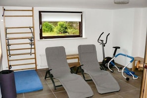 Ein Zimmer mit Fenster, eine Klimmzugstange, zwei Trainingsstühle, ein Ergometer und eine Trainingsmatte.