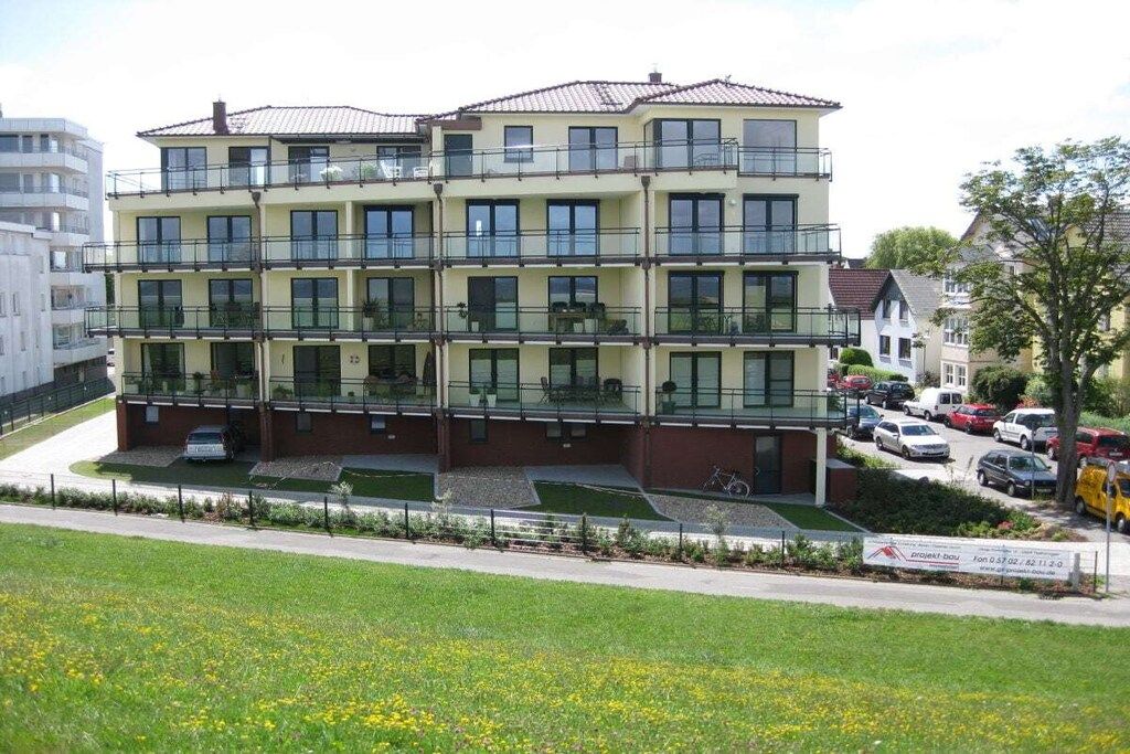 A multi-story residential building with balconies, a parking area, and a grassy field in the foreground.