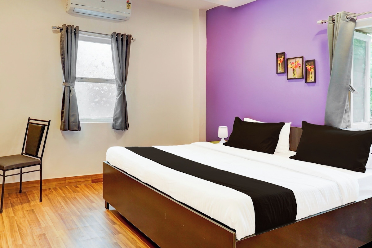 A bedroom with a bed, a chair, a window with curtains, and a wall with framed pictures.