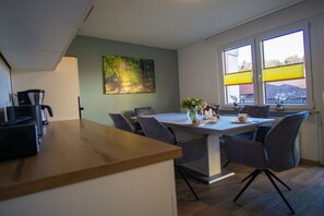 Un coin repas avec une table et des chaises, un tableau accroché au mur et une fenêtre avec des stores.