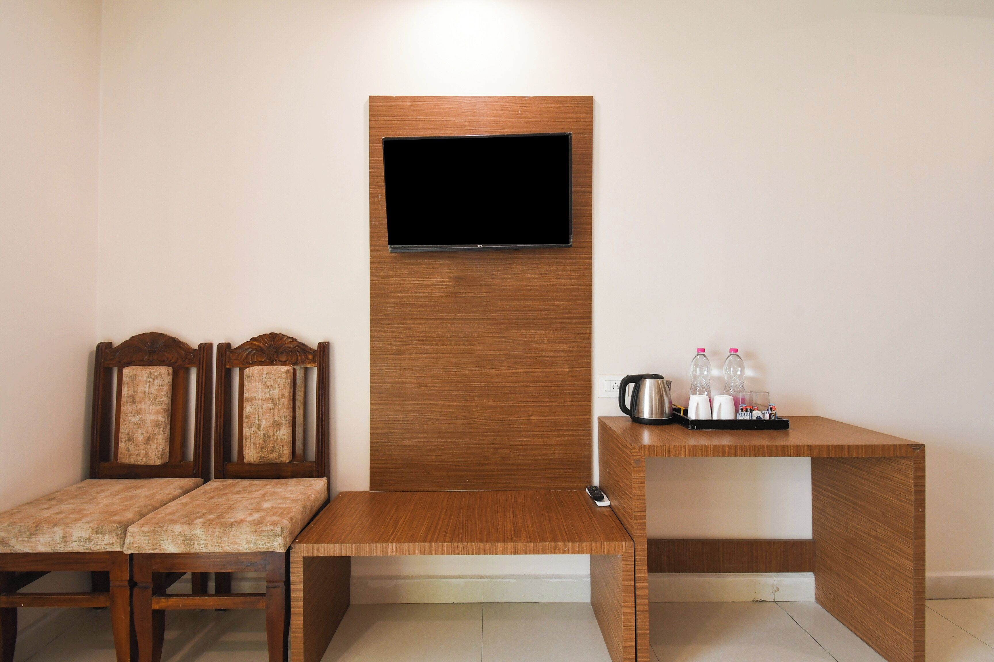 A room with a wooden bench, two upholstered chairs, a television mounted on a wooden panel, and a small table with a kettle and bottles.