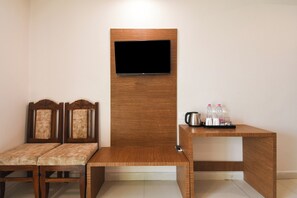 Quarto com um banco de madeira, duas poltronas estofadas, uma televisão fixada em um painel de madeira e uma mesinha com chaleira e garrafas.
