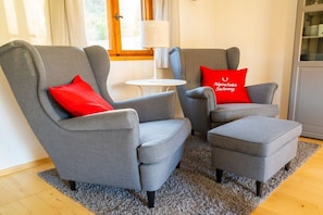 Een moderne zithoek met twee grijze fauteuils en een bijpassend voetenbankje, een klein wit bijzettafeltje en een rode kussen met tekst.