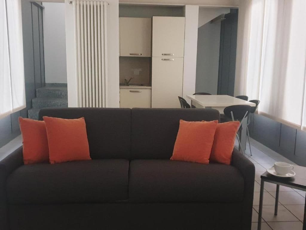 Un espacio de sala con un sofá y una mesa con sillas. Se observa un área de cocina con armarios y un refrigerador al fondo.